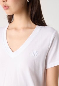 Vit bomullströja med v-ringning och korta ärmar. Har en broderad DKNY-logotyp i det övre vänstra hörnet. Mjuk textur.