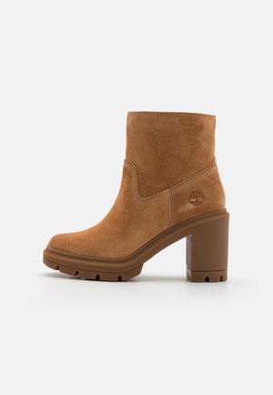 Tan suede ankle boot with a block heel, featuring a slightly rounded toe and stitching details. Logo embossed on the outer side.