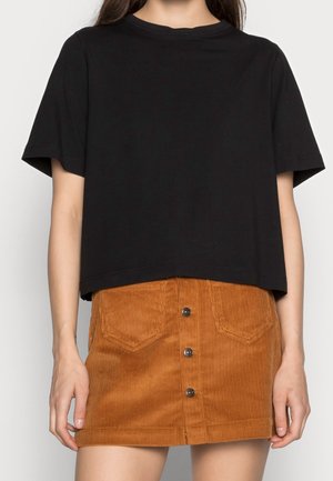 Kobieta w luźnej czarnej koszulce z krótkim rękawem, włożonej w wysoką brązową mini spódnicę z sztruksu zapinaną na guziki z przodu.