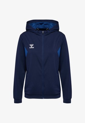 Granatowa bluza z kapturem na zamek błyskawiczny z regulowanymi troczkami, jaśniejsze niebieskie boczne panele, przednie kieszenie i białe logo hummel na lewej piersi.