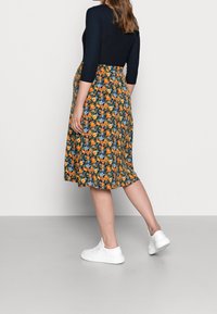 Květinová midi sukně v tmavě zelené barvě s oranžovými a modrými květy, vykazuje rozšířený tvar a hladký, lehký materiál. V kombinaci s bílými teniskami.