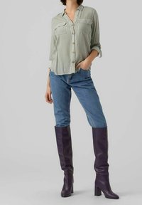 Kobieta w zielono-białej pionowo paskowanej bluzce, niebieskich dżinsach i wysokich fioletowych botkach na obcasie, stojąca z jedną ręką w kieszeni.