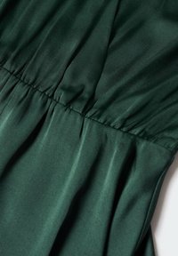 Tissu vert foncé avec une couture élastique froncée créant des plis subtils et une texture, suggérant un détail de vêtement ou de rembourrage.