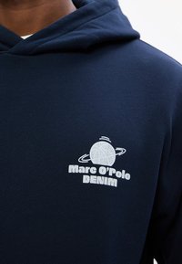 Hanorac bleumarin dintr-un material moale, cu un logo grafic alb ce prezintă un glob și inele, alături de textul "Marc O'Polo DENIM."