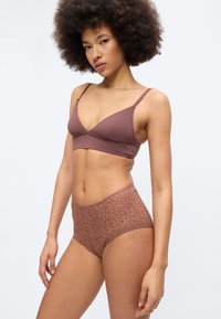 Γυναίκα με αφρό μαλλί που φοράει ένα μωβ μπραλέτα και καφέ εσώρουχο με λεοπάρ σχέδιο και ψηλή μέση, στέκεται μπροστά σε ένα απλό φόντο.