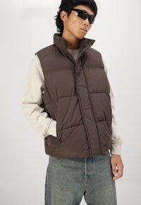 Brun polstret vest med høj krave, trykknapper og sidelommer. Båret over en beige langærmet skjorte med ribbede manchetter. Jeanser i denim til stede.