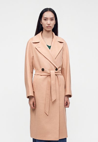 Femme portant un long manteau beige ceinturé avec des revers, debout droite devant un fond clair uni.