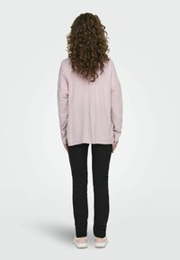 Chemise longue à manches longues rose clair, coupe décontractée, associée à un pantalon noir et des chaussures claires. Cheveux bouclés visibles de l'arrière.