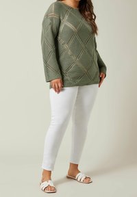 Kobieta w zielonym swetrze z dzianinowym wzorem w diamenty, białych dopasowanych spodniach i białych sandałach typu open-toe na płaskim obcasie, na neutralnym tle.