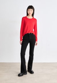 Κόκκινο μακρυμάνικο μπλουζάκι, εφαρμοστά μαύρα τζιν με καμπάνα και χοντρές μαύρες πλατφόρμες σε ένα μοντέλο μπροστά από ένα ουδέτερο φόντο.