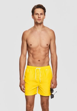 Młody mężczyzna stoi bez koszulki, ubrany w jaskrawożółte kąpielówki, trzyma czarne okulary przeciwsłoneczne w prawej ręce na jasnoszarym tle.