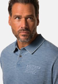 Světle modrá polo košile vyrobená z měkké látky, s límečkem, knoflíkovou légou a vyšitým textem "WESTERN RODEO SUPPLY" na hrudi.