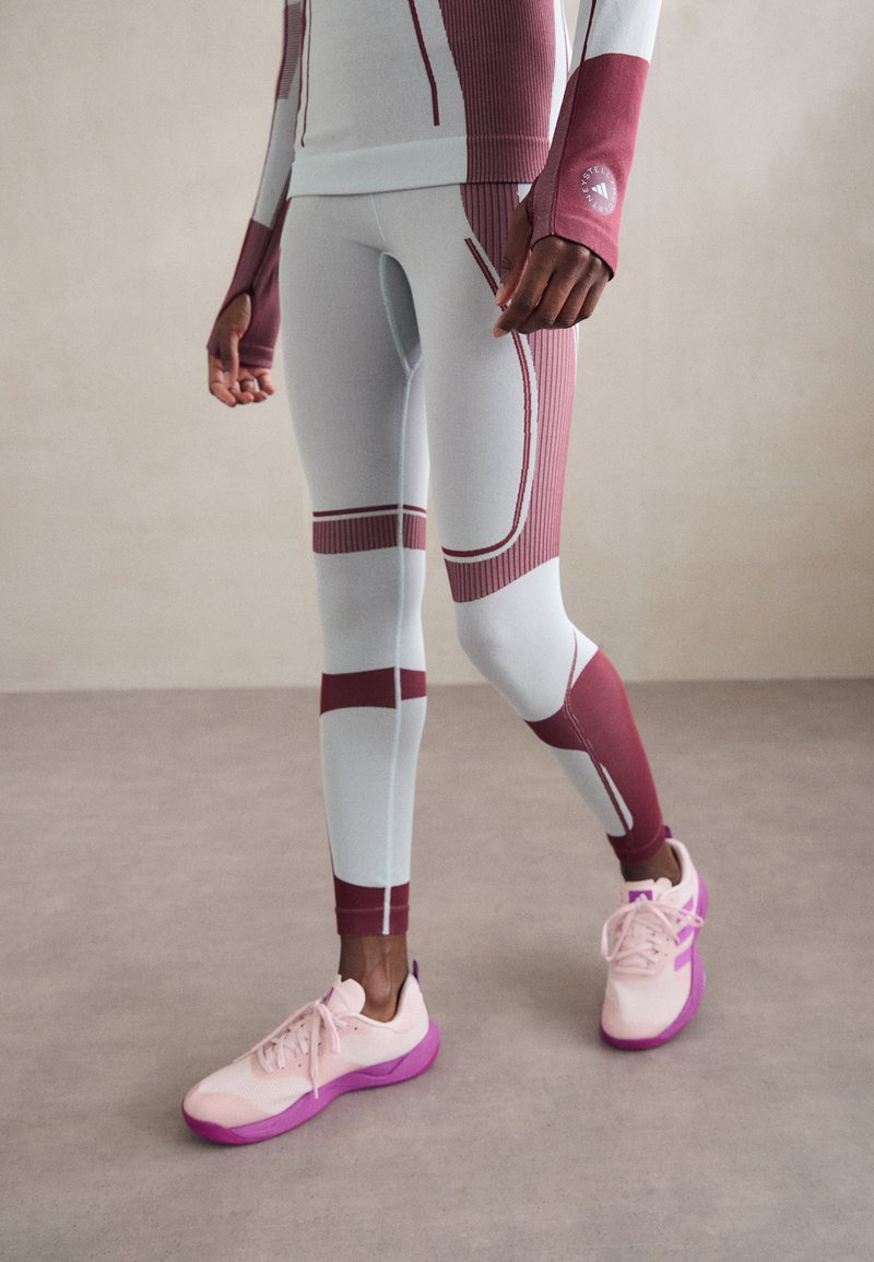 Személy fehér és bordó sport leggingsben és hozzáillő hosszú ujjú felsőben, rózsaszín és lila sportcipőben, áll egy szürke padlón.
