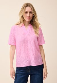 Blusa rosa a maniche corte con scollo a V e motivo a onde strutturate, realizzata in materiale leggero, abbinata a jeans blu scuro.