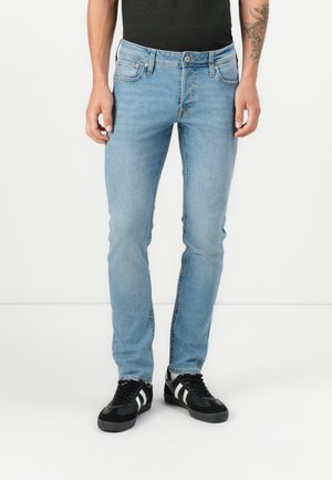 Homme portant un jean slim bleu clair et des baskets noires avec des rayures blanches, debout sur fond blanc.