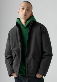 Ung mand med tætshavet hoved iført en grøn hættetrøje under en sort zip-up jakke med lommer, hænderne i jeanslommer, neutral baggrund.
