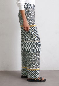 Un pantalon à jambes larges avec un design à motifs en bleu marine, blanc et or. Le tissu est léger, avec des motifs géométriques et une taille unie.