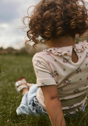 Dziecko o kręconych włosach siedzi na trawie, nosząc kwiecistą koszulę z falbankowym kołnierzem i zapięciem na guziki z tyłu, zwrócone tyłem do aparatu.