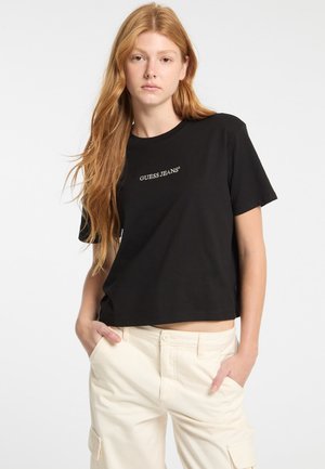 Čierne skrátené tričko vyrobené z mäkkého bavlneného materiálu s krátkymi rukávmi. Na prednej strane obsahuje biele logo "GUESS JEANS". Hladká textúra.