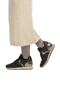 Schwarze und metallische Sneaker mit einem texturierten Obermaterial aus Stoff, braunen Lederakzenten und einer beigen Sohle, getragen mit hellgrauen gerippten Socken.