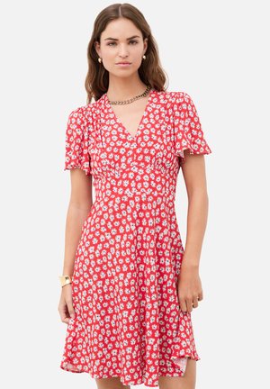 Femme portant une robe rouge à manches courtes avec imprimé marguerite blanche, col en V, et un collier en chaîne dorée ainsi qu'un bracelet sur un fond uni.