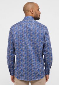 Blauwe blouse met lange mouwen, versierd met een bloemenpatroon in tinten blauw, goud en room. Gemaakt van gladde stof met een klassieke kraag en knopenmanchetten.