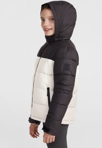 Giacca imbottita con cappuccio e maniche nere; corpo crema; presenta tasche con zip e una toppa con logo sulla manica. Materiale liscio e leggero.