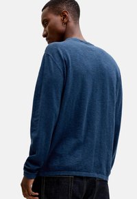 Homme portant une chemise bleue à manches longues et un jean foncé, debout de côté avec la tête légèrement inclinée vers le bas, devant un fond uni.