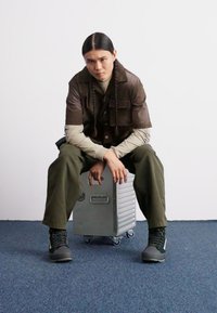 Caja de almacenamiento de metal plateado con superficie texturizada, apoyada sobre ruedas. Persona con camiseta marrón y pantalones verdes apoya las manos en la caja. Fondo neutro.