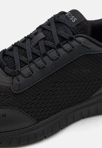 Svart atletisk sko med en strukturerad mesh-oberdel, gummisula och platta snören med en logodetalj på den vadderade tungan.