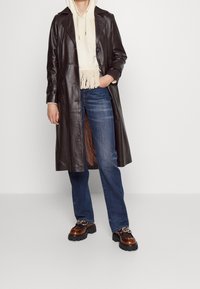 Un abrigo largo de cuero marrón con cierre de botones, sudadera beige con dobladillo con flecos, vaqueros azules y zapatos marrones de tacón grueso con detalles de herrajes.