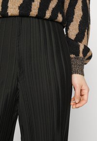 Calças pregueadas pretas com uma textura suave, combinadas com um suéter estampado em preto e castanho com punhos canelados.