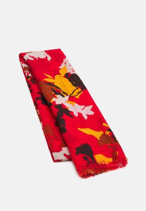 Gevouwen rood stof met levendige gele, witte, zwarte en bruine bloemmotieven en rafelige randen op een witte achtergrond.