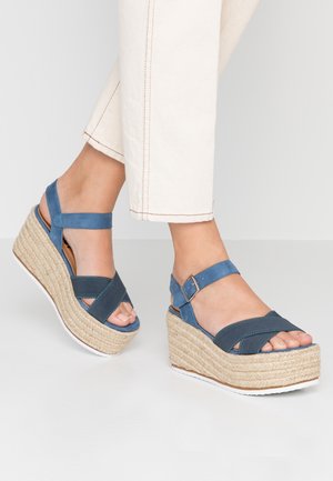Pieds portant des sandales compensées bleues avec brides à la cheville et un pantalon beige court, sur un fond blanc uni.