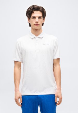 Νέος άνδρας με σκούρα μαλλιά, φορώντας λευκό πόλο μπλουζάκι Calvin Klein και μπλε παντελόνι με διακριτικά αφηρημένα σχέδια, στέκεται μπροστά σε απλό φόντο.