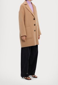 Abrigo de lana color beige con cuello con muescas, tres botones y bolsillos de parche, combinado con jeans de pierna ancha negros y sandalias negras.