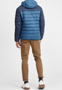 Blå og marineblå puffet jakke med hætte, med quiltet tekstur. Parret med brune bukser og hvide sneakers. Set fra ryggen.
