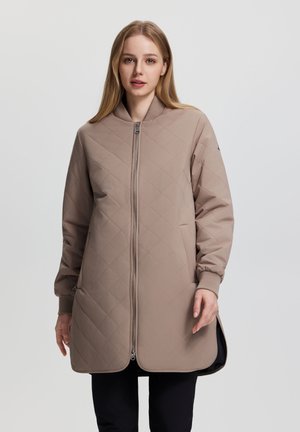 Junge Frau mit glatten blonden Haaren trägt eine lange, beige gesteppte Jacke mit Reißverschluss vorne und steht vor einem einfarbigen grauen Hintergrund.