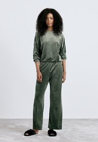 Vrouw met krullend haar die staat, gekleed in een groene fluwelen lounge-set en zwarte fuzzy pantoffels op een lichtgrijze vloer.