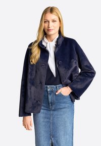 Jachetă din blană sintetică bleumarin cu guler, având două nasturi și o croială lejeră, asortată cu o fustă din denim bleu deschis și o bluză albă.