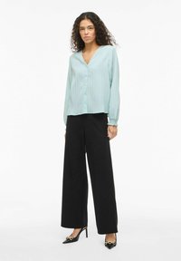 Gai zila plīša blūze ar V veida izgriezumu un pogām, komplektā ar augsta vidukļa melnām, platu kāju biksēm un melniem apaviem ar smailiem purngaliem.