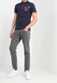 Polo shirt blu navale con accenti rossi e bianchi, caratterizzato da una chiusura con tre bottoni e un logo. Abbinato a pantaloni grigi e sneakers bianche.