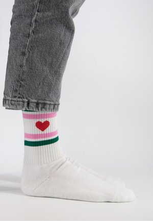Noga w szarych dżinsach, na stopie białe skarpetki z różowo-zielonymi paskami i czerwonym sercem, stojąca na jednolitym białym tle.