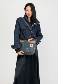 Giacca di jeans blu con colletto alto e tasche anteriori, abbinata a una lunga gonna nera. Tieni in mano una borsa di pelle blu con motivi e accenti.