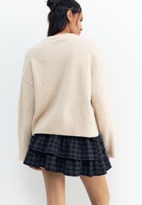 Femeie cu păr lung și închis la culoare, purtând un pulover bej și o fustă în carouri bleumarin stratificată, cu fața întoarsă spre un fundal alb simplu.