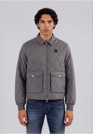 Junger Mann mit lockigem dunklem Haar trägt eine graue gesteppte Jacke mit zwei Vordertaschen und blaue Jeans, steht vor einem schlichten grauen Hintergrund.