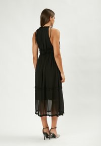 Vestido de gasa negra con escote halter, cintura fruncida y acentos de volantes en capas. Presenta un diseño hasta la rodilla y una textura transparente.
