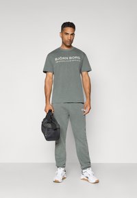 Pelēks kokvilnas T-krekls ar uzsistu tekstu "BJÖRN BORG TRAINING CLUB MONACO", apvienots ar saskaņotām treniņbiksēm, melnu sporta somu un baltām kedām.