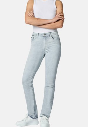 Frau trägt hellblaue Slim-Fit-Jeans, weißes ärmelloses Oberteil und weiße Sneaker, steht mit verschränkten Armen vor einfarbigem Hintergrund.