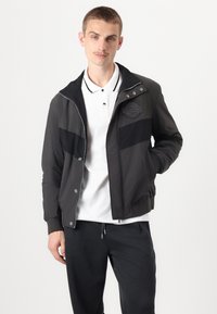 Giacca bomber nera in nylon con chiusura a zip e bottoni a pressione, caratterizzata da una patch del logo e strisce a contrasto, abbinata a una polo bianca.
